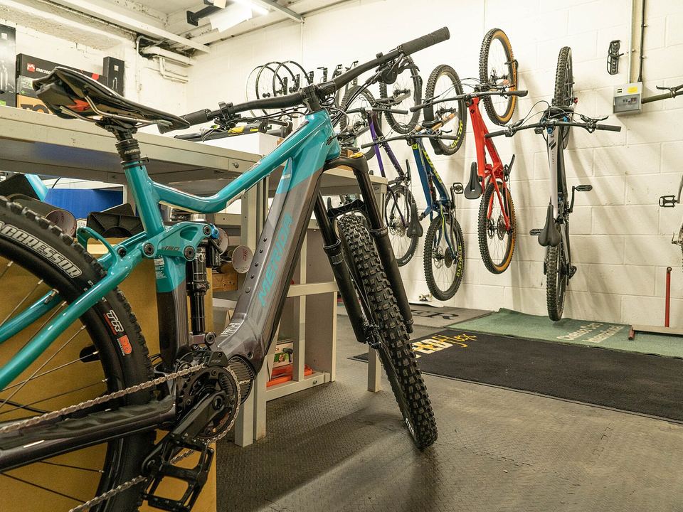 A teal electric mountain bike in the workshop, with more bikes hanging on the white brick wall behind it